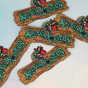 Könnte beinhalten: Gestickte Aufnäher mit dem Schriftzug "Splash Mountain" in Türkis auf braunem Holzmaserung-Hintergrund. Jeder Aufnäher zeigt ein Cartoon-Kaninchen mit einer roten Erdbeere. Geeignet für Jacken, Taschen oder andere Textilien.