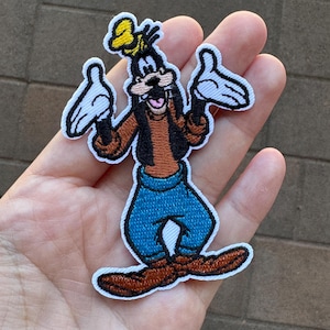 May include: Embroidered iron-on patch featuring a cartoon image of Goofy, a Disney character, with his signature goofy grin and outstretched arms. He is wearing a brown shirt, blue jeans, and brown shoes.
