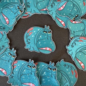 Op de afbeelding: Een verzameling teal geborduurde patches met een cartoon nijlpaard met een humeurige uitdrukking. Het nijlpaard heeft een roze mond, rode accenten en gele wimpers. De patches zijn verspreid over een donkere ondergrond.