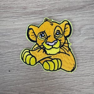 Peut inclure: Un patch brodé jaune à repasser représentant un lionceau de dessin animé avec de grands yeux bleus. Le lionceau est assis, les pattes croisées, et a un nez rose.