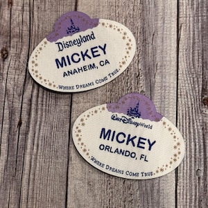 Może przedstawiać: Dwa owalne identyfikatory z fioletowym zamkiem i złotymi gwiazdami. Górny identyfikator zawiera napis "Disneyland MICKEY ANAHEIM, CA WHERE DREAMS COME TRUE". Dolny identyfikator zawiera napis "WALT Disney World MICKEY ORLANDO, FL WHERE DREAMS COME TRUE".