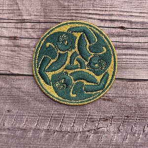 Może przedstawiać: Okrągła, zielono-złota naszywka z haftem przedstawiającym celtycki węzeł. Węzeł ma kształt potrójnej spirali.