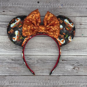 May include: A pair of black mouse ears with a brown and orange sequined bow. The ears are decorated with a print of a cartoon scene with the text "Wildest Ride" repeated on each ear.