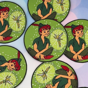Könnte beinhalten: Runde, gestickte Aufnäher mit Peter Pan und Tinkerbell. Peter Pan trägt eine grüne Tunika und einen Hut, rotes Haar und rote Schuhe. Tinkerbell trägt ein gelbes Kleid mit weißen Flügeln. Der Hintergrund ist grün mit weißen Punkten.