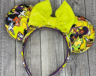 Op de afbeelding: Gele en paarse stoffen muisoren met een gele strik en een cartoon-personageontwerp met Goofy, Pluto en andere Disney-personages.