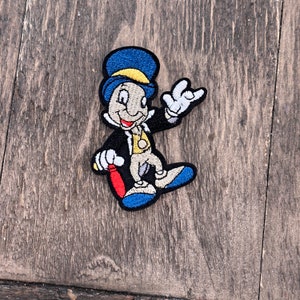 Könnte beinhalten: Ein gestickter Aufnäher mit einer Cartoonfigur mit einem blauen Hut, einer gelben Weste und einem roten Stock. Die Figur lächelt und hat einen schelmischen Ausdruck.