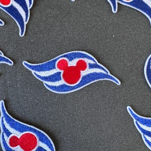 May include: Embroidered patches with a blue and white design and a red Mickey Mouse head. The patches have a wing-like shape and are set against a grey background. These patches are suitable for adding to clothing or accessories.