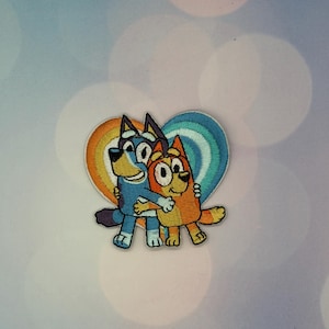 May include: An embroidered patch featuring two cartoon dogs, one blue and one orange, standing in front of a heart-shaped background with rainbow-colored stripes. The dogs are embracing each other, with the blue dog on the left and the orange dog on the right, both depicted with simple outlines and stitched details.