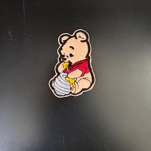 May include: Embroidered iron-on patch featuring Winnie the Pooh, a cartoon bear, in a red shirt, holding a honey pot and dripping honey.