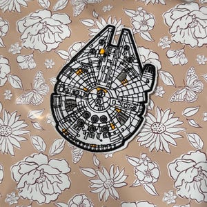 Pode incluir: Um remendo bordado branco e preto do Millennium Falcon, a nave espacial de Star Wars. O remendo está sobre um fundo marrom com um padrão floral.