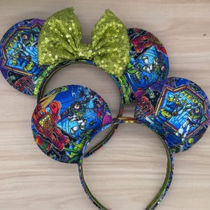May include: Three headbands with round ears featuring a Buzz Lightyear design. The ears are blue with a colorful print of Buzz Lightyear and other characters. A green sequined bow sits atop one headband.
