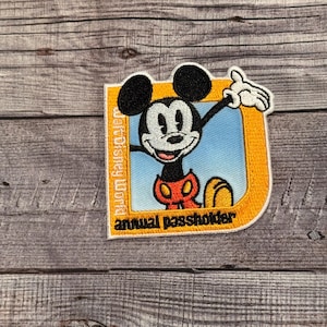 以下が含まれることがあります： ミッキーマウスが描かれた黄色と青の刺繍パッチ。テキストは「Walt Disney World annual passholder」です。