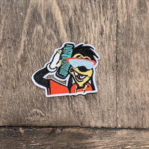 Op de afbeelding: Een geborduurde strijk-op patch met een cartoon hond die een zonnebril en een gouden ketting draagt. De hond houdt een blauwe en groene kam in zijn rechterpoot. De patch is wit met oranje, gele, zwarte en blauwe accenten.