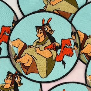 May include: Embroidered patch featuring a cartoon character in shades of brown, orange, and green, set against a light blue circular background. The patch has a black border and depicts a character with a llama-like appearance.