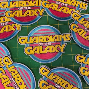 May include: Embroidered patches featuring the text "GUARDIANS OF THE GALAXY" in yellow, outlined in white, on a pink and blue circular background. The patches are arranged on a green grid.