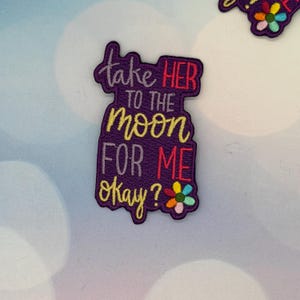 Puede incluir: Un parche bordado morado con el texto "take HER to the moon FOR ME okay?" en blanco, amarillo y rojo. Una pequeña flor con pétalos coloridos está en la parte inferior derecha. El parche es un accesorio único.