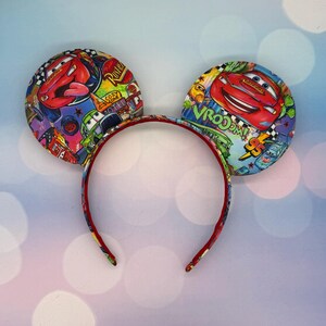 May include: Mickey Mouse ear headband featuring a colorful Cars movie design. The ears display images of Lightning McQueen and other characters, with a red headband. The ears are round and padded, with a vibrant, cartoonish aesthetic.