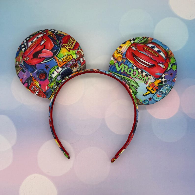 May include: Mickey Mouse ear headband featuring a colorful Cars movie design. The ears display images of Lightning McQueen and other characters, with a red headband. The ears are round and padded, with a vibrant, cartoonish aesthetic.
