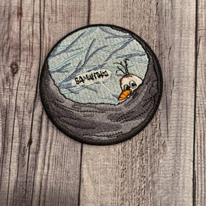 以下が含まれることがあります： 氷の穴から顔を出している雪だるまが刺繍されたパッチ。パッチは青い背景に白い線が入り、"Samantha"という文字が書かれている。