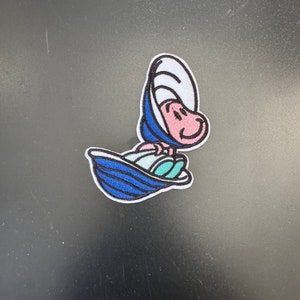 May include: An embroidered patch featuring a cartoon character in a blue and white shell, riding a blue and white wave. The character has a pink face and a happy expression.