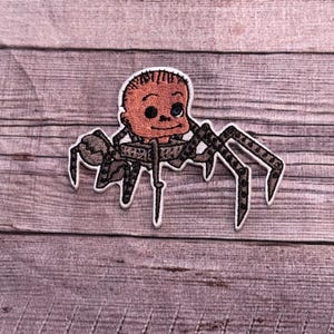 May include: Embroidered patch featuring a cartoon baby face with a spider body. The baby face is peach-colored with black eyes and a small smile. The spider body is brown with black accents and eight legs.