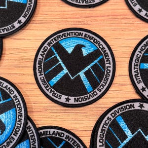 May include: Circular black and blue embroidered patches with the text "STRATEGIC HOMELAND INTERVENTION ENFORCEMENT LOGISTICS DIVISION" and a stylized eagle emblem. Patches are scattered on a light brown wooden surface.