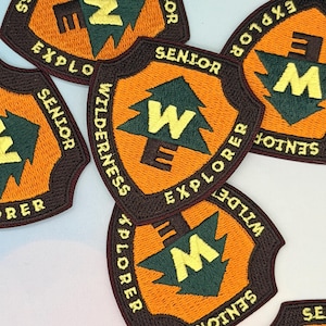 May include: Embroidered patches with a shield design. The patches feature the text "SENIOR EXPLORER WILDERNESS" in brown, with a green tree and the letters "W" and "E" in yellow. The background is orange.