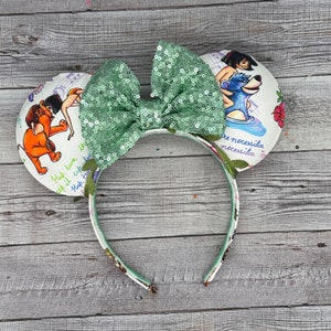 May include: Headband with two round ears featuring illustrations of characters from The Jungle Book, including Mowgli, Baloo, and an elephant. The ears have a white background with green text and a large, central sage green sequined bow with leaves.