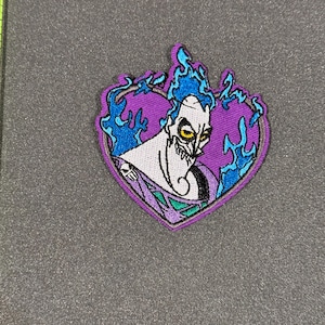 May include: Purple heart-shaped embroidered patch featuring Hades from Disney's Hercules. Hades is depicted with blue flames and a menacing expression.