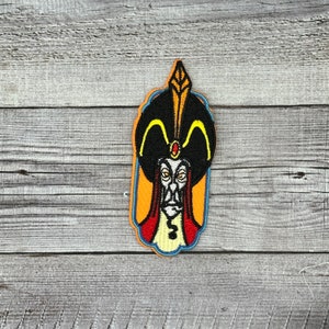 May include: An embroidered patch featuring a cartoon depiction of Jafar from Disney's Aladdin. Jafar is wearing a black turban with a yellow jewel and a red robe. He has a long, white beard and a sinister expression.
