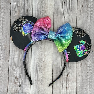 May include: Black mouse ear headband with a rainbow sequin bow. The ears feature fireworks and a Main Street Electrical Parade logo on one ear and a neon dragon on the other.