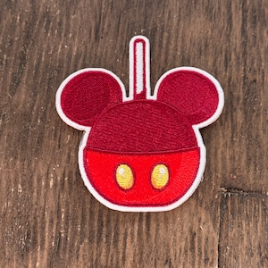 May include: Red and white embroidered patch of a Mickey Mouse head with a red candy apple body and yellow eyes.