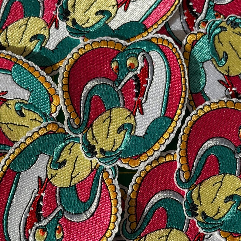 May include: A collection of embroidered patches featuring a stylized snake design. The patches have a red heart-shaped background, a green snake, and a yellow skull. The snake has a white belly and a red tongue. The patches are outlined with a gold border.