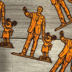 May include: Orange embroidered patches featuring a man in a suit and Mickey Mouse. The man is waving, and Mickey Mouse is holding up his hand. The patches are on a wooden surface.