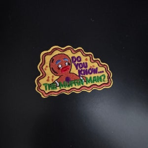 Peut inclure: Un écusson brodé jaune et marron avec un personnage de bonhomme en pain d'épices. Le bonhomme en pain d'épices a un bouton violet et dit "Connais-tu... le vendeur de muffins ?"