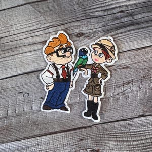 Könnte beinhalten: Stickersatz mit gestickten Figuren aus dem Animationsfilm Obendrauf. Die Figuren sind ein Mann mit Brille und eine Frau mit Hut, die beide einen grünen Vogel halten.