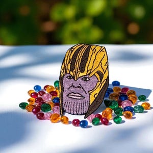 May include: Embroidered patch featuring the face of Thanos, with a purple face and gold helmet. The patch is surrounded by colorful glass beads in shades of red, orange, green, blue, and pink. The background is blurred.
