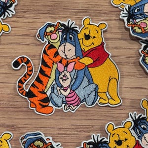 Puede incluir: Parche bordado con Winnie de Pooh, Tigger, Piglet y Eeyore. Los personajes están todos sonriendo y abrazándose. El parche está hecho de fieltro y tiene un contorno blanco.
