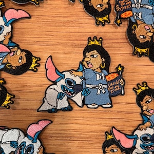 Puede incluir: Parches bordados con un personaje de dibujos animados con un vestido azul y una corona, sosteniendo una bolsa de caramelos, con una criatura blanca y azul. Los parches tienen detalles en rosa, azul y dorado. El fondo es una superficie de madera marrón claro.