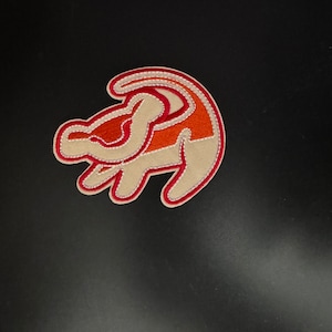Puede incluir: Parche bordado con la silueta de una cabeza de león en naranja, beige y rojo. El parche tiene un diseño detallado con un borde rojo, adecuado para aplicar en ropa o accesorios.