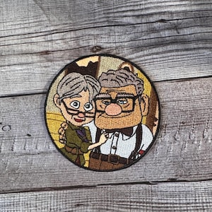 Op de afbeelding: Gestikte patch met een cartoon-koppel, een man met bruin haar en een vrouw met grijs haar, die elkaar omhelzen. De man draagt een wit overhemd, een strikje en bretels. De vrouw draagt een groene jurk. De patch heeft een bruine achtergrond met een geel huis op de achtergrond.