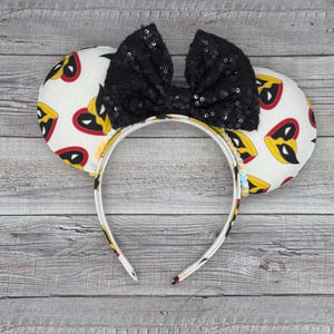 May include: Black sequin bow headband with white fabric mouse ears featuring a repeated pattern of yellow and red superhero masks.