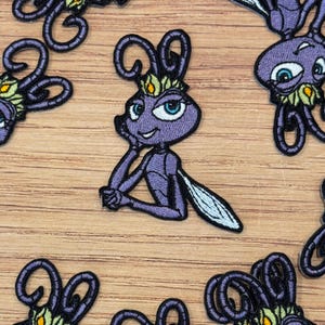 Op de afbeelding: Geborduurde patch met een paars insectenpersonage met grote ogen, een kroon en een bedachtzame pose. De patch heeft een gedetailleerd ontwerp met zwarte contouren en accenten van blauw, wit en geel. De patches zijn gerangschikt op een houten oppervlak.