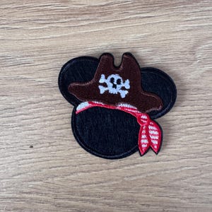 May include: A black and brown embroidered patch of a cartoon character wearing a pirate hat with a skull and crossbones design. The character is wearing a red and white striped bandana.