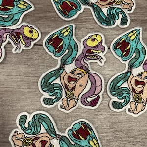 May include: Embroidered patches featuring a cartoon character with a baby face, held by green and purple snakes. The patches have a white border and are set against a light wood grain background.
