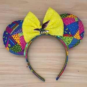 May include: A colorful headband with mouse ears. The ears are patterned with a patchwork design in various colors. A large yellow sequined bow is centered on the headband, adorned with a bird embellishment. The headband is designed for wear.