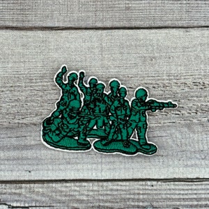 May include: Green plastic army men figures in a group, standing and holding weapons. The figures are arranged in a line, with some facing forward and others facing sideways. The figures are all wearing helmets and have a cartoonish appearance.