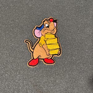 May include: An embroidered patch of a brown mouse carrying a stack of yellow cheese. The mouse is wearing a yellow shirt, a pink and blue hat, and red shoes.