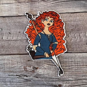 Może przedstawiać: Naszywka haftowana do prasowania przedstawiająca postać z kreskówki z długimi czerwonymi włosami trzymającą łuk i strzałę. Postać ma na sobie niebieską tunikę i brązowe skórzane spodnie.