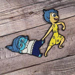May include: Two embroidered patches featuring characters from the animated film Inside Out. The patch on the left is blue and depicts Sadness, a character with blue skin, short blue hair, and glasses. The patch on the right is yellow and depicts Joy, a character with yellow skin, long blue hair, and a yellow dress with a sun pattern.